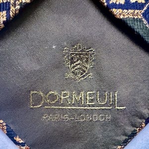 May include: A necktie with a dark gray lining featuring an embroidered crest and the text DORMEUIL PARIS-LONDON. The tie fabric displays a geometric pattern in shades of blue, green, and gold with intricate leaf-like motifs throughout.