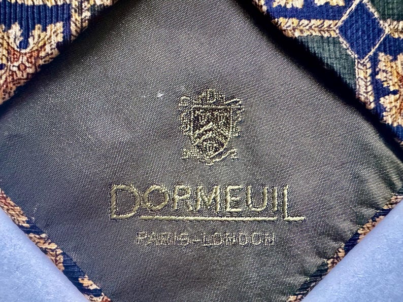 May include: A necktie with a dark gray lining featuring an embroidered crest and the text DORMEUIL PARIS-LONDON. The tie fabric displays a geometric pattern in shades of blue, green, and gold with intricate leaf-like motifs throughout.