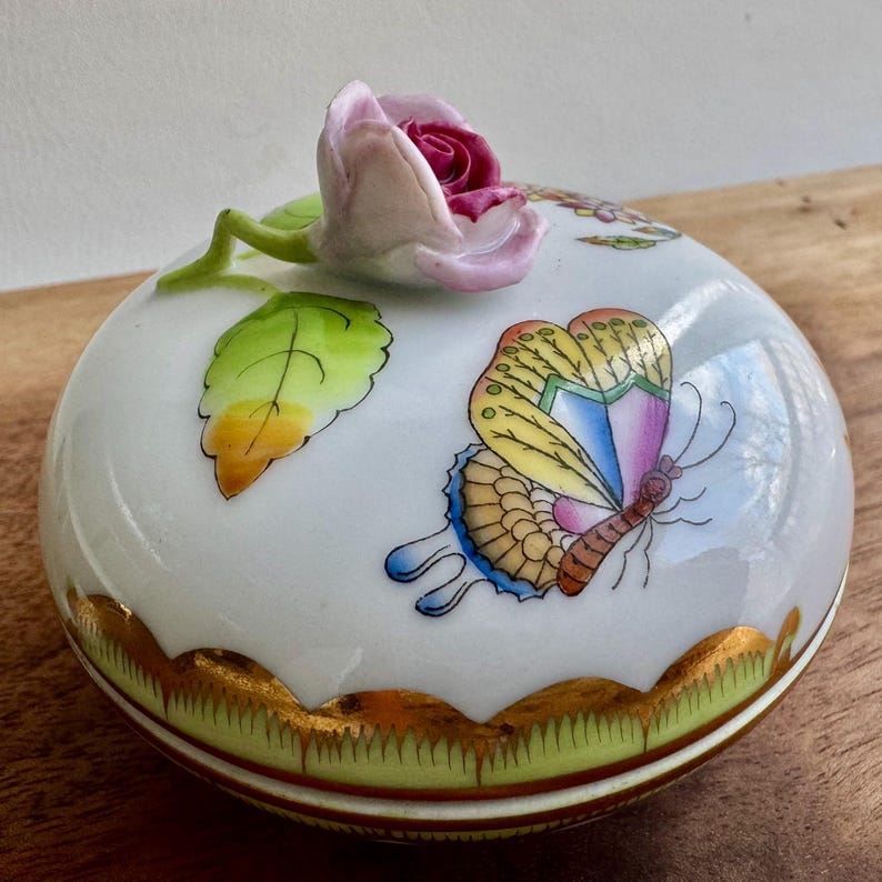 May include: A white porcelain trinket box with a gold rim and a green and gold base. The lid features a hand-painted butterfly with yellow, orange, pink, and blue wings. A pink rose with green leaves sits on top of the lid.