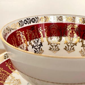 Pode incluir: Close-up de uma ch&aacute;vena e pires brancos com uma faixa vermelha e um design de filigrana dourada. A ch&aacute;vena tem uma forma arredondada com uma borda dourada. O pires tem um design a condizer. O tema geral &eacute; a loi&ccedil;a vintage.