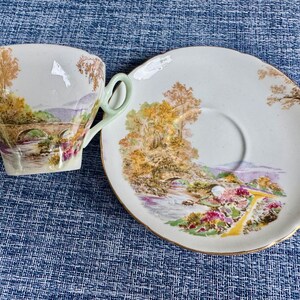 May include: A white porcelain teacup and saucer set featuring a colorful landscape illustration of a stone bridge over a river, surrounded by trees and flowers. The teacup has a pale green handle, and both pieces have scalloped edges with gold trim.