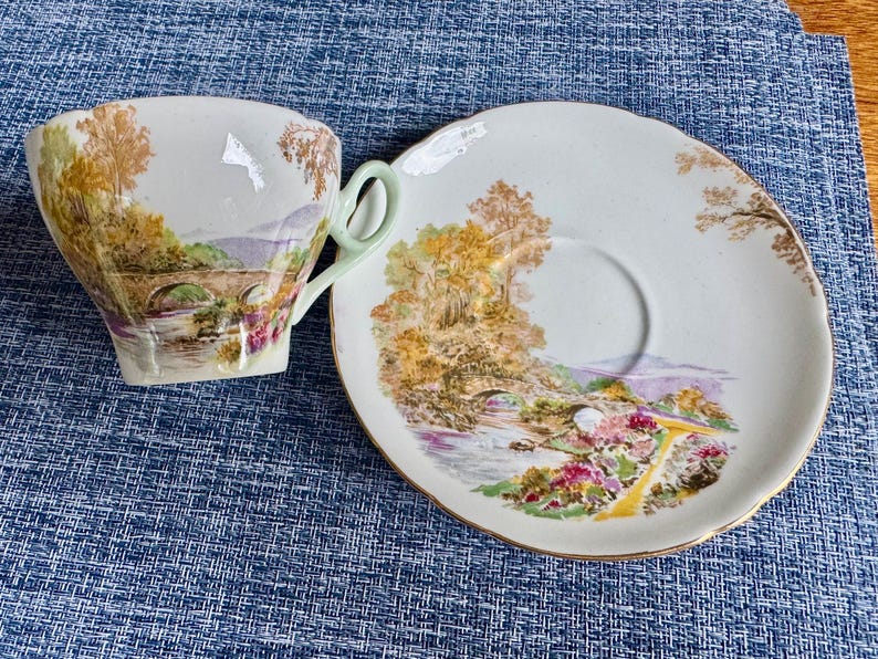 May include: A white porcelain teacup and saucer set featuring a colorful landscape illustration of a stone bridge over a river, surrounded by trees and flowers. The teacup has a pale green handle, and both pieces have scalloped edges with gold trim.