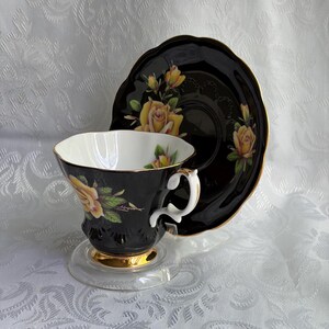 Pode incluir: Um conjunto de ch&aacute;vena e pires em porcelana preta com padr&otilde;es de rosas amarelas, bordas recortadas douradas e interior branco. A ch&aacute;vena tem uma asa branca curva com detalhes dourados e uma base dourada, sobre um pires de cor preta.