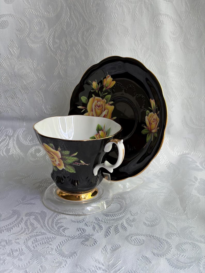 Pode incluir: Um conjunto de ch&aacute;vena e pires em porcelana preta com padr&otilde;es de rosas amarelas, bordas recortadas douradas e interior branco. A ch&aacute;vena tem uma asa branca curva com detalhes dourados e uma base dourada, sobre um pires de cor preta.