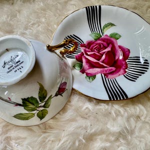 May include: A white teacup and saucer set with a floral rose design. The saucer features a large pink rose with green leaves and black and white stripes. The teacup has a gold handle and a rose and leaf design.