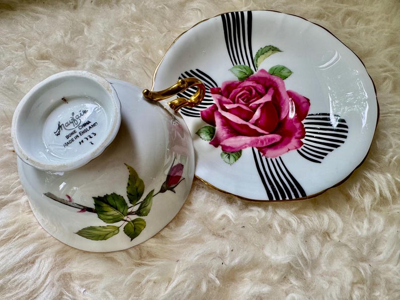 May include: A white teacup and saucer set with a floral rose design. The saucer features a large pink rose with green leaves and black and white stripes. The teacup has a gold handle and a rose and leaf design.