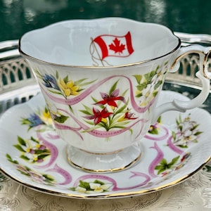 May include: White teacup and saucer set with gold trim, featuring floral and ribbon designs. The teacup has a Canadian flag and the names of Canadian provinces. The saucer and cup have a scalloped edge.