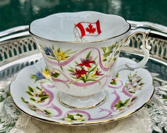 May include: White teacup and saucer set with gold trim, featuring floral and ribbon designs. The teacup has a Canadian flag and the names of Canadian provinces. The saucer and cup have a scalloped edge.