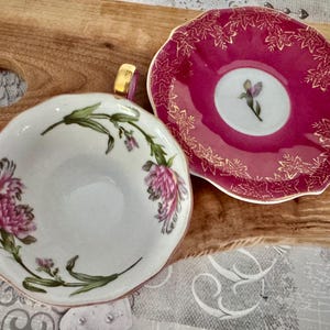 May include: A white teacup and saucer set with floral designs. The teacup features pink flowers and green leaves, while the saucer is a deep pink with gold leaf accents. The handle of the teacup is gold.