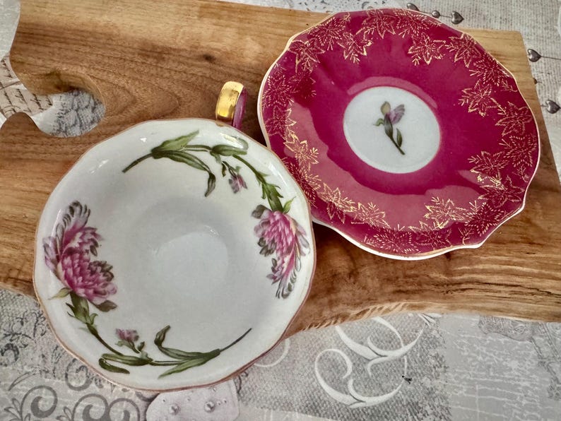 May include: A white teacup and saucer set with floral designs. The teacup features pink flowers and green leaves, while the saucer is a deep pink with gold leaf accents. The handle of the teacup is gold.