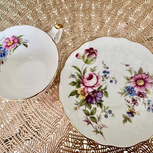 May include: A white teacup and saucer set with a floral design. The cup and saucer have a gold trim and feature a variety of pink, purple, blue, and yellow flowers.