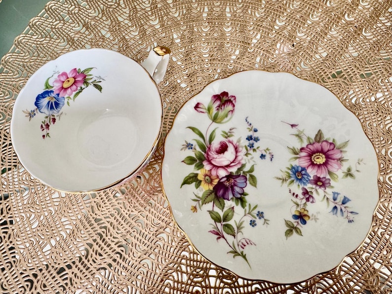 May include: A white teacup and saucer set with a floral design. The cup and saucer have a gold trim and feature a variety of pink, purple, blue, and yellow flowers.