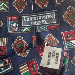 May include: A navy blue necktie featuring a repeating pattern of small rectangular crests and diamond shapes in red, green, and white. A black label reads CHRISTOPHER BARNES, and a white tag indicates 100% SILK / SOIE and CA00125.