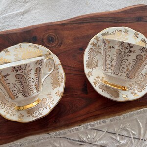 May include: Two white teacups and saucers with gold floral patterns. The cups have a delicate, feminine design.