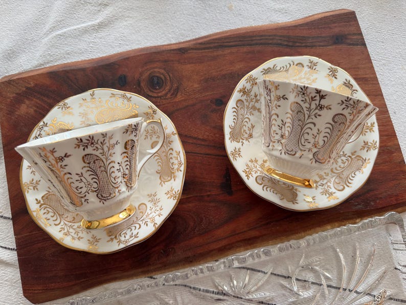 May include: Two white teacups and saucers with gold floral patterns. The cups have a delicate, feminine design.