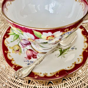 May include: A vintage teacup and saucer set with a floral design. The teacup and saucer have a white background with red trim and gold accents. A silver spoon rests on the saucer. The teacup and saucer are placed on a gold-colored surface.