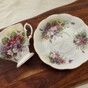 May include: A white teacup and saucer set with a floral design featuring purple, pink, and white flowers. The flowers are surrounded by green leaves and a yellow background.