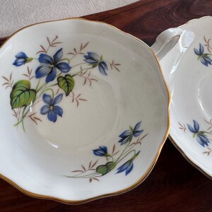 May include: Two white porcelain teacups with a gold rim and a floral design featuring blue and green violets.