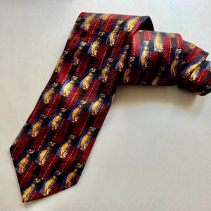 May include: A necktie featuring a repeating pattern of yellow golf bags on blue squares, separated by vertical bands of textured dark red fabric. The tie has a pointed end and is folded over itself, displaying the intricate woven design throughout.