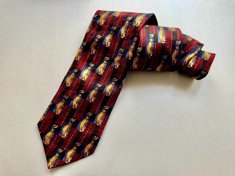 May include: A necktie featuring a repeating pattern of yellow golf bags on blue squares, separated by vertical bands of textured dark red fabric. The tie has a pointed end and is folded over itself, displaying the intricate woven design throughout.