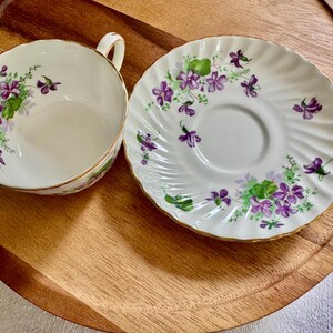 K&ouml;nnte beinhalten: Ein wei&szlig;es Tassen- und Untertassen-Set mit floralem Design. Die Tasse hat einen Henkel und einen goldenen Rand. Die Untertasse hat einen gewellten Rand und ein passendes Blumenmuster. Die Blumen sind lila und gr&uuml;n.
