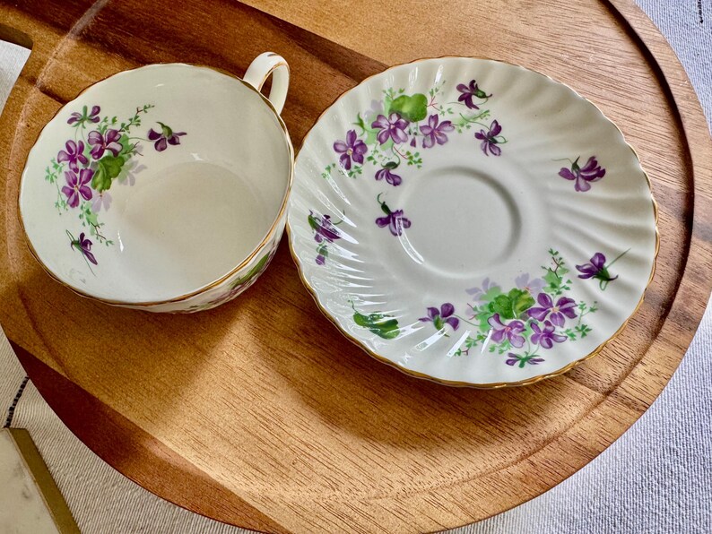 K&ouml;nnte beinhalten: Ein wei&szlig;es Tassen- und Untertassen-Set mit floralem Design. Die Tasse hat einen Henkel und einen goldenen Rand. Die Untertasse hat einen gewellten Rand und ein passendes Blumenmuster. Die Blumen sind lila und gr&uuml;n.