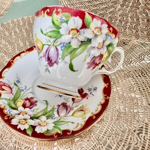May include: A white teacup and saucer set with a red border and gold trim. The cup and saucer are decorated with colorful floral designs, including white, purple, and yellow flowers with green leaves. The set is placed on a gold-colored surface.