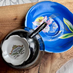 Puede incluir: Un juego de taza y platillo de t&eacute; en blanco y negro con un dise&ntilde;o floral. El platillo es azul con un borde dorado y una flor blanca con acentos amarillos y rosados. La taza de t&eacute; tiene un borde negro y un interior blanco con las palabras "Princess China Hand Occupied Japan" impresas en la parte inferior.