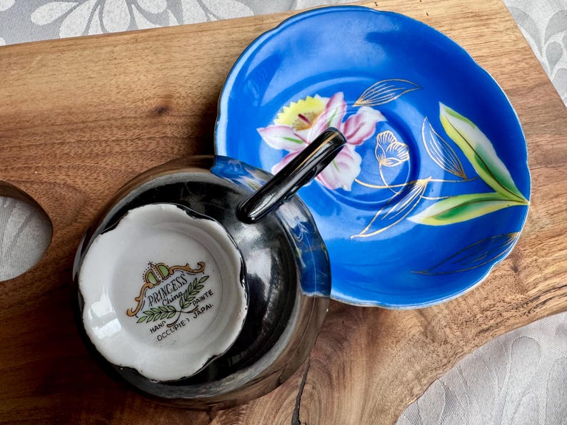 Puede incluir: Un juego de taza y platillo de t&eacute; en blanco y negro con un dise&ntilde;o floral. El platillo es azul con un borde dorado y una flor blanca con acentos amarillos y rosados. La taza de t&eacute; tiene un borde negro y un interior blanco con las palabras "Princess China Hand Occupied Japan" impresas en la parte inferior.