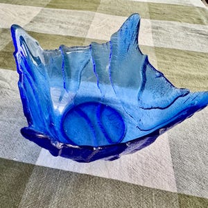 Pu&ograve; includere: Una ciotola in vetro blu cobalto vibrante con una forma organica irregolare. La ciotola presenta una superficie ruvida e ondulata che imita l'aspetto dell'acqua o del ghiaccio, con bordi alti e appuntiti e una base circolare liscia.
