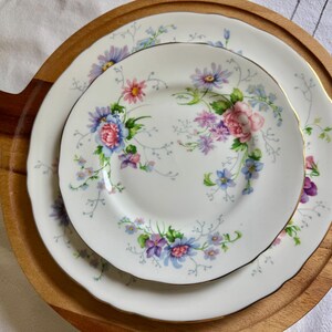May include: White porcelain plates with a floral pattern in pink, blue, and purple, presented on a round wooden serving tray. The plates have gold edging and a scalloped rim, ideal for serving food.
