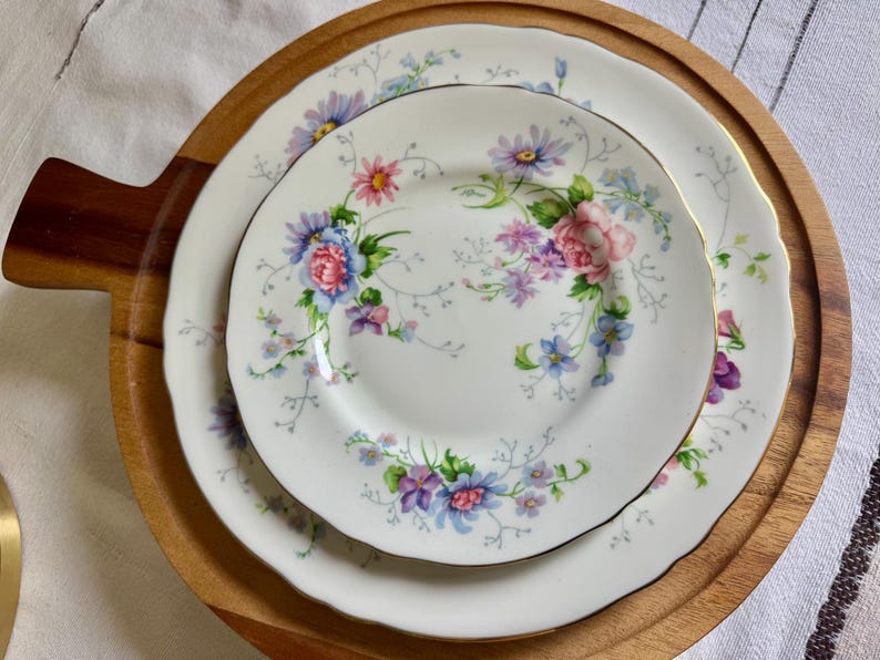 May include: White porcelain plates with a floral pattern in pink, blue, and purple, presented on a round wooden serving tray. The plates have gold edging and a scalloped rim, ideal for serving food.