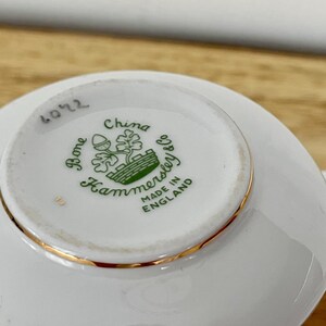 May include: A white bone china teacup and saucer set. The underside of the cup features a green and gold Hammersley & Co. logo with the words "Bone China" and "Made in England". The rim is trimmed with gold.