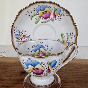 May include: A white teacup and saucer set with a floral pansy design in blue, yellow, and purple. The rims are trimmed with gold. The teacup sits on a clear glass stand.
