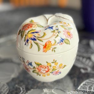 K&ouml;nnte beinhalten: Eine wei&szlig;e Porzellan-Schmuckdose in Apfelform, verziert mit einem floralen Muster in den Farben Blau, Gelb, Rot und Rosa.