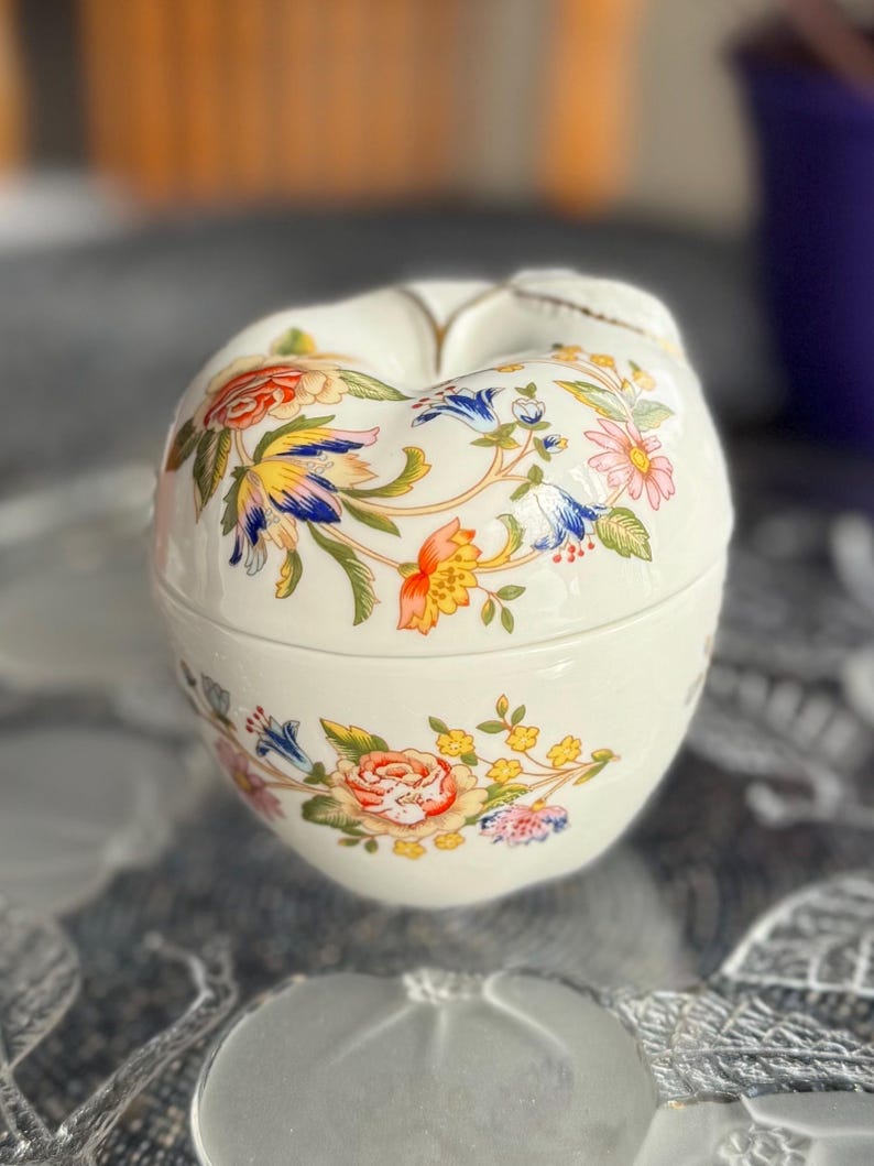 K&ouml;nnte beinhalten: Eine wei&szlig;e Porzellan-Schmuckdose in Apfelform, verziert mit einem floralen Muster in den Farben Blau, Gelb, Rot und Rosa.
