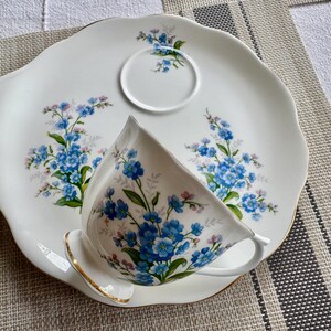 May include: A white porcelain teacup and saucer set with a forget-me-not floral pattern. The set is on a white and gray striped placemat.