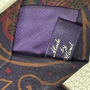 Pode incluir: Uma gravata com base roxo-escura e um padr&atilde;o paisley multicolorido em tons de castanho, vermelho e verde. A parte de tr&aacute;s da gravata inclui um forro de tecido roxo e uma pequena etiqueta retangular com o texto Made by Hand.