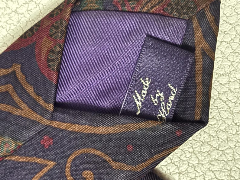 Pode incluir: Uma gravata com base roxo-escura e um padr&atilde;o paisley multicolorido em tons de castanho, vermelho e verde. A parte de tr&aacute;s da gravata inclui um forro de tecido roxo e uma pequena etiqueta retangular com o texto Made by Hand.