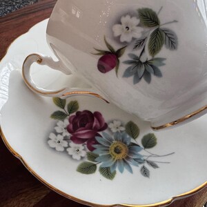 May include: A white teacup and saucer set with a gold trim. The teacup features a floral design with red, white, and blue flowers. The saucer has a similar floral design with a larger blue flower.