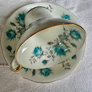 Peut inclure: Un service &agrave; th&eacute; vintage compos&eacute; d'une tasse et d'une soucoupe. La porcelaine blanc cass&eacute; pr&eacute;sente des motifs floraux de roses turquoise avec des feuilles grises et une bordure dor&eacute;e.