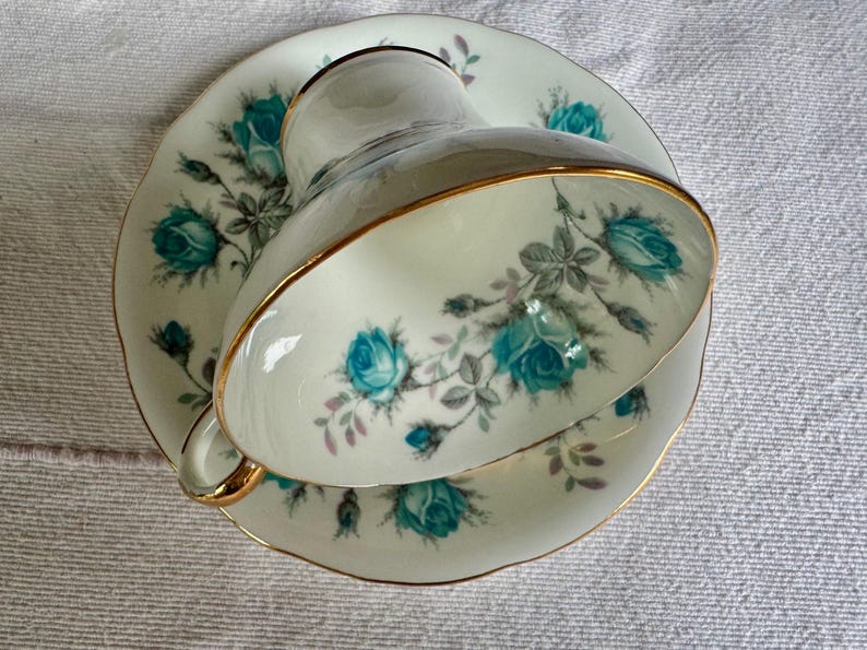 Peut inclure: Un service &agrave; th&eacute; vintage compos&eacute; d'une tasse et d'une soucoupe. La porcelaine blanc cass&eacute; pr&eacute;sente des motifs floraux de roses turquoise avec des feuilles grises et une bordure dor&eacute;e.