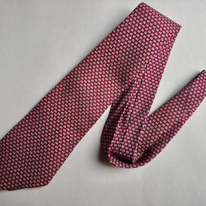 Puede incluir: Una corbata con fondo rojo intenso y un patr&oacute;n repetitivo de peque&ntilde;as formas geom&eacute;tricas claras. La corbata est&aacute; doblada, mostrando el extremo puntiagudo y la secci&oacute;n m&aacute;s estrecha, con una textura consistente en todo el tejido.