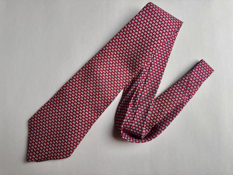 Puede incluir: Una corbata con fondo rojo intenso y un patr&oacute;n repetitivo de peque&ntilde;as formas geom&eacute;tricas claras. La corbata est&aacute; doblada, mostrando el extremo puntiagudo y la secci&oacute;n m&aacute;s estrecha, con una textura consistente en todo el tejido.