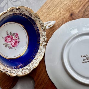 May include: A vintage teacup and saucer set with a blue and gold floral design. The teacup has a white interior with a pink rose pattern. The saucer has a white interior with the words "Royal Stafford Bone China Made in England" printed in gold.