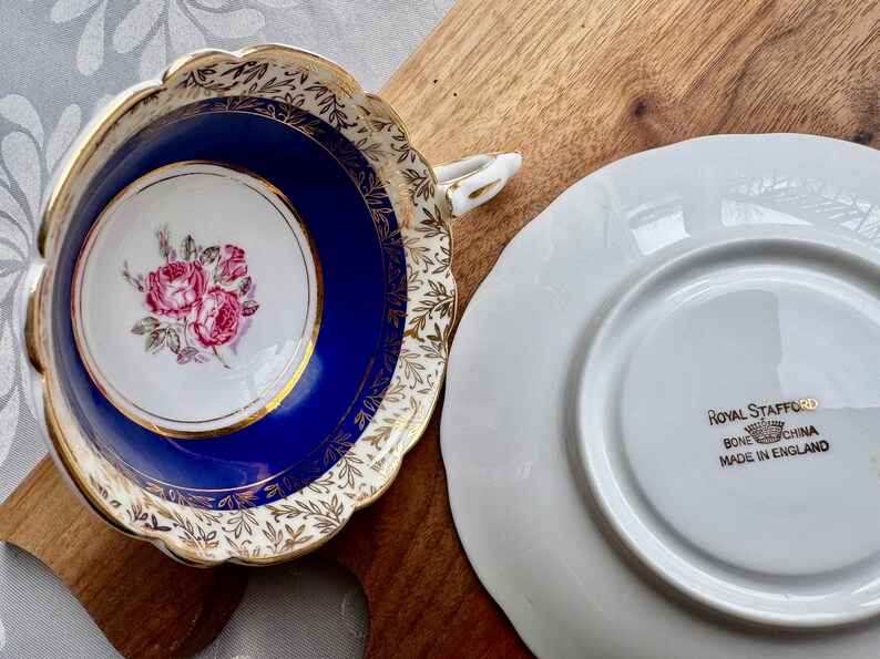 May include: A vintage teacup and saucer set with a blue and gold floral design. The teacup has a white interior with a pink rose pattern. The saucer has a white interior with the words "Royal Stafford Bone China Made in England" printed in gold.