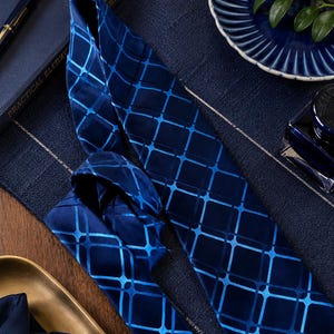 May include: A navy blue tie with a geometric grid pattern in a lighter shade of blue. The tie is displayed on a textured navy blue surface, alongside a matching pocket square. A pen and a book are visible in the background.