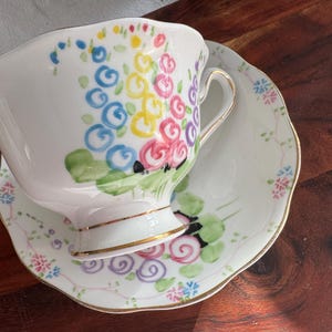 May include: A white teacup and saucer with a floral design. The cup and saucer are decorated with hand-painted flowers in shades of pink, blue, yellow, and green.