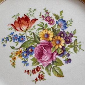 May include: White ceramic plate with a floral design. The arrangement features a large pink rose, a red tulip, and various other flowers in shades of yellow, blue, and purple. The rim has a gold and blue border.
