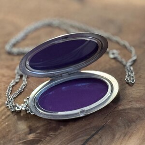 Puede incluir: Collar con medall&oacute;n ovalado plateado, abierto para revelar un interior morado oscuro. El medall&oacute;n est&aacute; sobre una cadena plateada, sobre una superficie de madera. El medall&oacute;n tiene un dise&ntilde;o ornamentado, con una est&eacute;tica cl&aacute;sica y vintage.